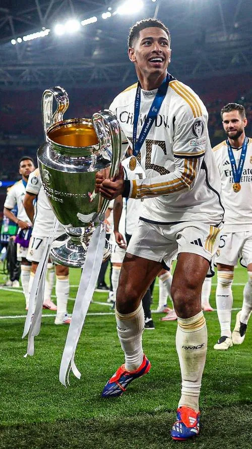 1 Hình đẹp Jude nâng cúp Champions League, áo trắng nổi bật, ánh đèn sân làm gương mặt sáng rõ.