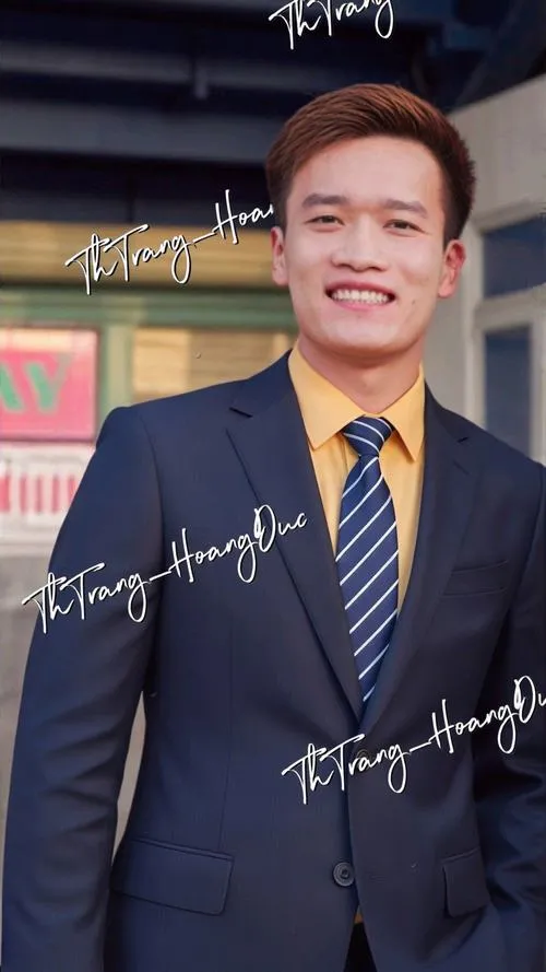 19 Hoàng Đức mặc vest dự sự kiện, cà vạt vàng, nụ cười tự tin, hậu cảnh banner và ánh đèn