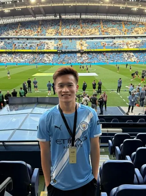 1 Hoàng Đức ngồi khán đài sân vận động, áo xanh, thẻ đeo, nền cỏ và khán đài xa