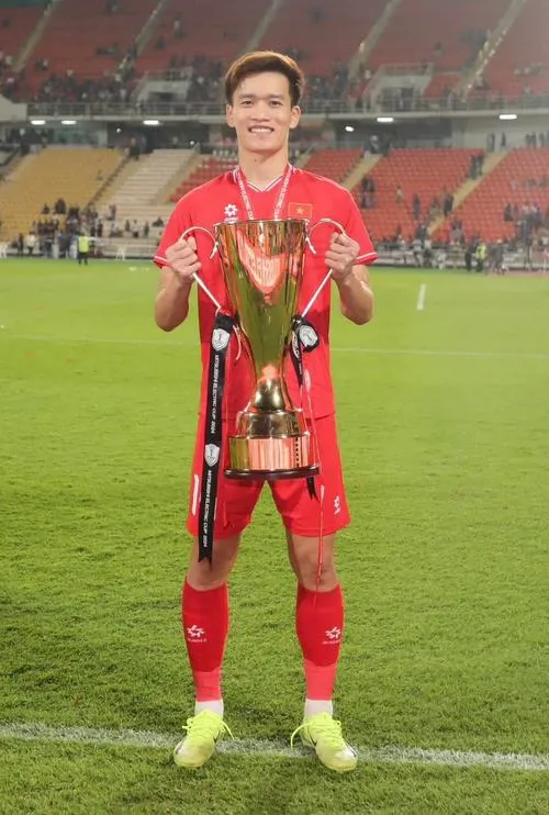 2 Cầu thủ Hoàng Đức áo đỏ trên sân cỏ, ôm cúp, dáng cao, ánh đèn sân vận động rực