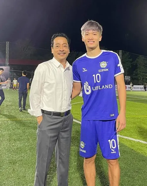 4 Hoàng Đức mặc áo xanh số 10 đứng cạnh người lớn tuổi, nụ cười nhẹ, sân cỏ phía sau