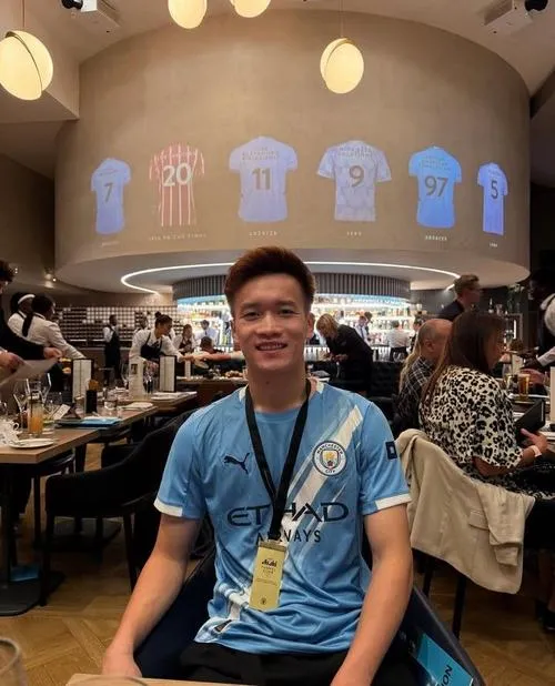 8 Hoàng Đức ngồi quán có áo đấu treo tường, áo xanh, dáng thoải mái, cảm giác gần gũi