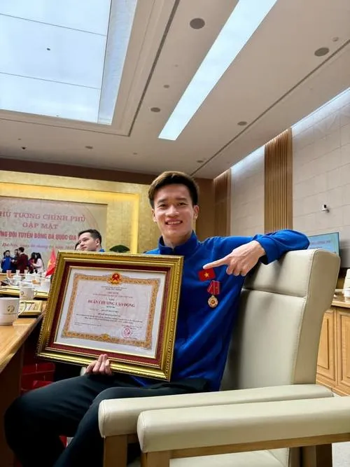 16 Hoàng Đức ngồi ghế sofa cầm bằng khen, cười hiền, bối cảnh phòng khách sáng và gọn