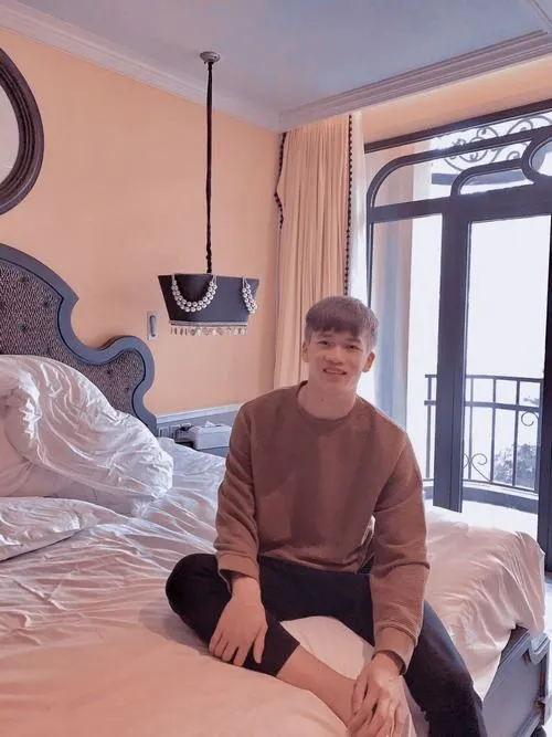 17 Hoàng Đức ngồi trên giường trong phòng, áo nâu, ánh sáng cửa sổ, vẻ trầm và bình yên