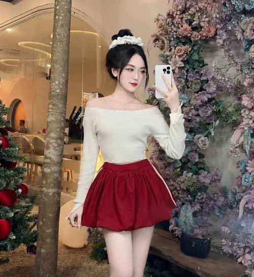 44 Cô gái phối áo trắng trễ vai cùng chân váy đỏ xếp ly đứng cạnh cụm cây và đèn vàng, bờ vai lộ nhẹ cùng nụ cười làm khung hình vừa dễ thương vừa quyến rũ một cách rất tự nhiên