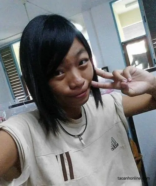 24 Cô gái mặc áo trắng giơ tay chữ V trong phòng học hoặc văn phòng, mái tóc chéo một bên và nụ cười mím khiến khung hình trông như ảnh hài hước pha chút duyên dáng, đơn giản mà vẫn có điểm nhấn riêng