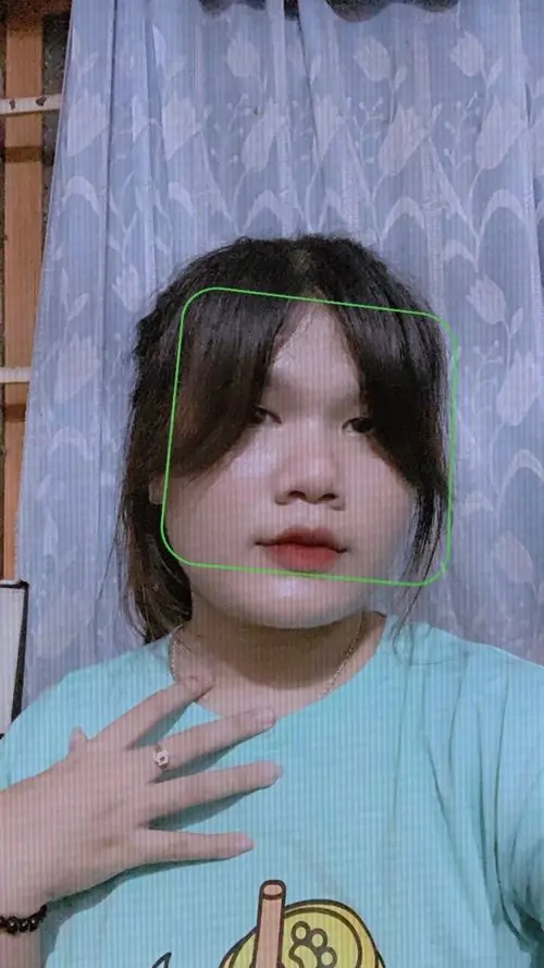 4 Bé gái áo xanh đứng cạnh rèm tím, một tay đặt trước ngực và mái tóc rủ che bớt mắt, bố cục dọc làm đường nét khuôn mặt trông mềm hơn, tạo cảm giác ảnh nữ chụp khoảnh khắc xấu nhưng vẫn rất đời thường