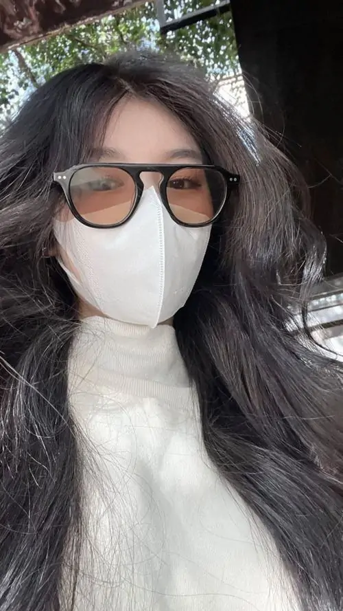 2 Cô gái đeo kính lớn, kéo cao cổ áo len trắng che nửa dưới gương mặt khi đứng ngoài gió, phần mắt sau tròng màu và tóc buông dài làm bức hình mang vẻ cuốn hút kín đáo, hiện đại, lạnh nhẹ mà vẫn sang