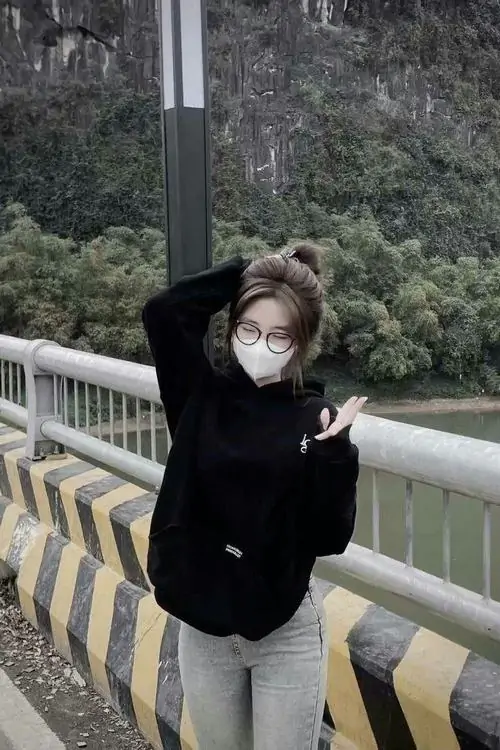 8 Cô gái mặc đồ đen trên lan can đường núi, đeo khẩu trang trắng che kín nửa mặt rồi nghiêng người giơ tay tạo dáng, khung cảnh ngoài trời giúp bức ảnh gái đẹp che mặt trở nên trẻ trung, gọn gàng và đầy năng lượng