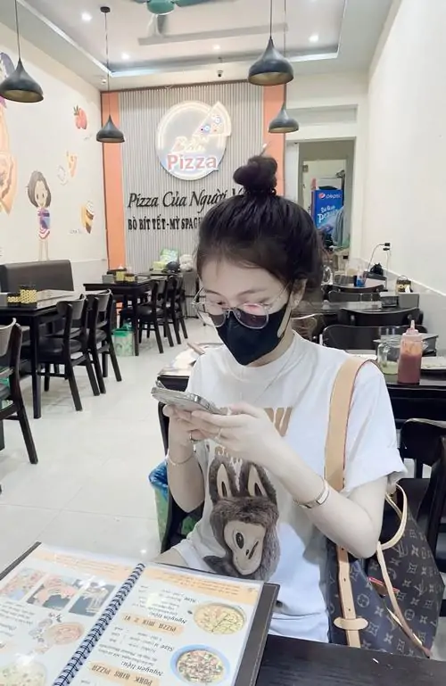 11 Hình gái xinh ngồi trong quán ăn sáng, đeo khẩu trang đen che phần dưới gương mặt rồi cúi nhìn tờ giấy trước mặt, tóc búi cao và outfit trắng đen tạo cảm giác gọn, sạch, có nét cá tính rất hợp ảnh đời thường