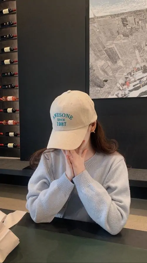 26 Cô gái ngồi trong quán, đội mũ be và chắp hai tay trước mặt như che gần kín nửa trên khuôn mặt, chỉ còn lộ cằm và tóc dài, bức ảnh mang cảm giác kín đáo, đáng yêu nhẹ nhưng vẫn giữ được độ cá tính riêng