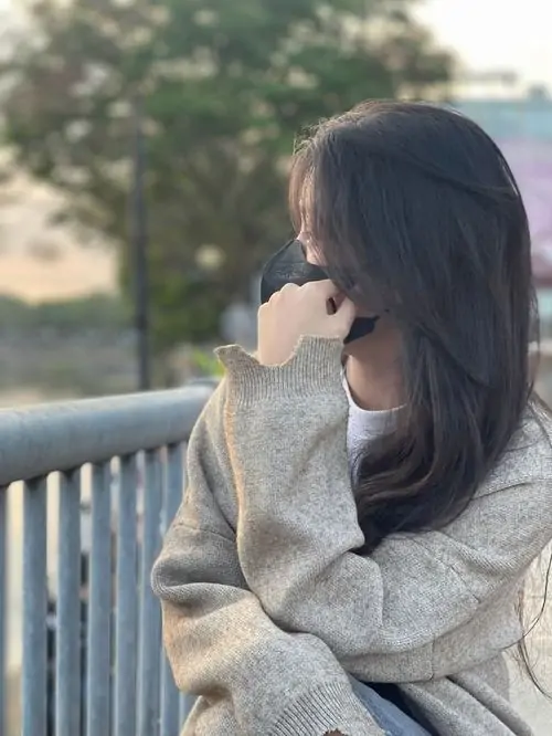 41 Hình nàng quay lưng đứng bên lan can trong áo len rộng, xoay người ra phía sáng để toàn bộ mặt khuất khỏi ống kính, mái tóc nâu và bối cảnh thoáng tạo một vẻ dịu hơn, nhưng chất ảnh vẫn giữ được độ lạnh cần có