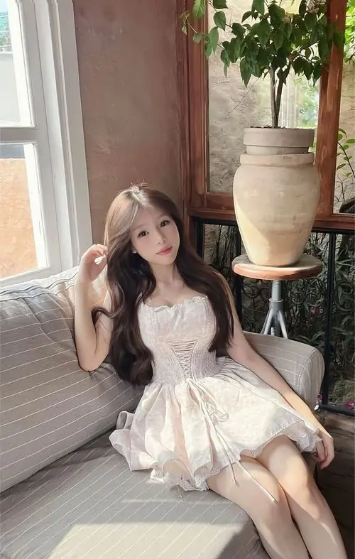 8 Ngồi trên ghế trong không gian nội thất sáng, cô gái mặc váy kem bồng nhẹ, tay đặt khéo và chân khép tự nhiên, bối cảnh gỗ cùng chậu cây làm khung hình mềm hơn, trông dễ thương như một nàng thơ