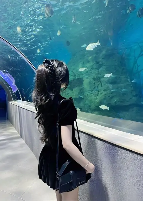 26 Cô gái mặc váy sẫm đứng trước bể cá quay lưng để đường tóc suôn dài nổi lên giữa nền xanh mát