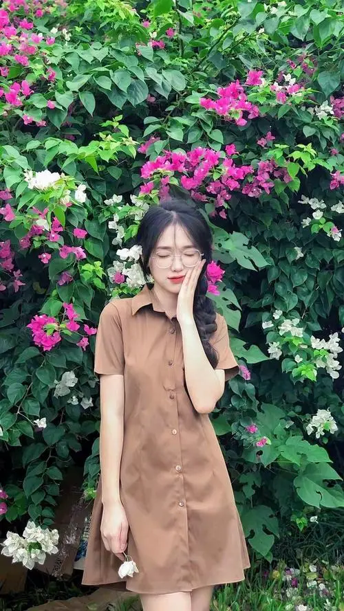 2 Gái đẹp đứng trước khóm hoa tím trong ảnh nữ sinh đeo kính xinh với váy nâu nhẹ và nụ cười mềm