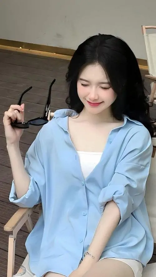 30 Cô gái ngồi với áo sơ mi xanh nhạt, tóc đen buông thẳng và nụ cười kín đáo, ảnh chân dung nữ cuốn hút theo kiểu hiền nhẹ vì cảm xúc truyền tải ấm áp và dáng ngồi gọn gàng khiến tổng thể rất đẹp