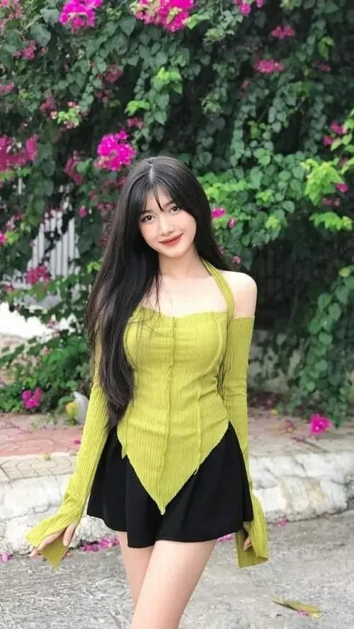 16 Cô gái mặc thiết kế vàng đen đứng trước giàn hoa tím, mái tóc dài phủ vai và dáng nghiêng khoe đường cong tạo cảm giác quyến rũ rõ rệt, đồng thời giữ được nét cute nhờ gương mặt mềm