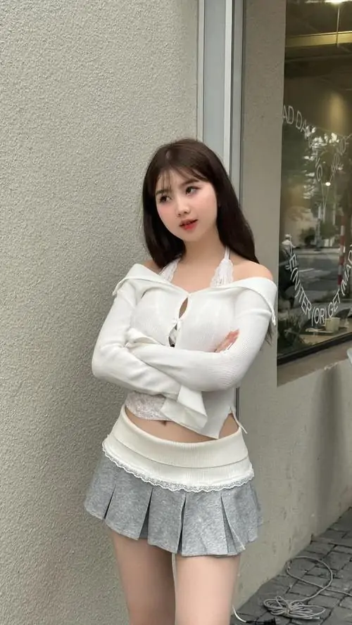 21 Khung ảnh cô gái mặc áo trắng tay dài cùng chân váy xếp ly xám đứng trong sảnh sáng, vòng tay ôm trước ngực làm dáng vẻ kín mà vẫn tôn thần thái, hợp kiểu ảnh nữ xinh trong váy