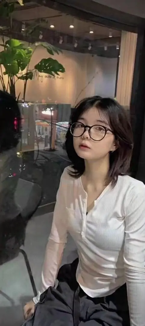 16 Hot girl đeo kính ngồi nghiêng trong quán với áo trắng ôm nhẹ và váy tối màu, mái tóc ngang vai cùng ánh mắt hướng ra ngoài tạo cảm giác suy tư dễ mến, khung hình vừa thanh lịch vừa mang nét cuốn hút riêng