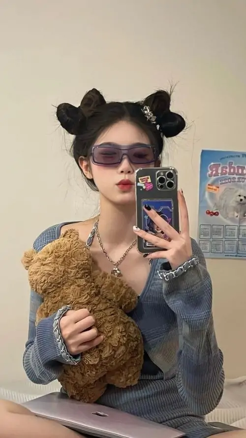 22 Cô nàng đeo kính tạo dáng selfie với kiểu tóc búi đôi và áo khoác xanh, tay ôm chú gấu bông lớn trước gương, nét đẹp cá tính đến từ biểu cảm bình thản cùng không gian phòng ngủ ấm áp và đáng yêu