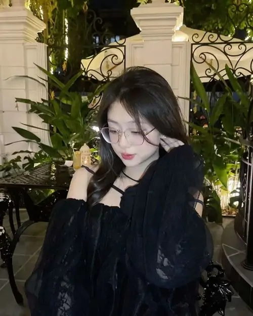 30 Gái xinh mang kính ngồi giữa không gian nhiều cây và ánh đèn vàng, áo khoác tối ôm dáng, mái tóc dài phủ xuống gương mặt, sắc lạnh hiện lên vừa đủ trong góc cúi đầu khiến bức ảnh đêm thêm sâu và nổi bật