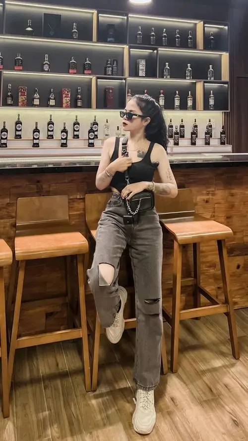 7 Khung hình nàng thơ chụp trong quán với kệ chai phía sau, cô gái mặc đồ tối màu ngồi trên ghế cao, chân dài thon gọn và tư thế thoải mái, gái xinh mang kính hiện lên sành điệu, nổi bật giữa không gian ấm áp