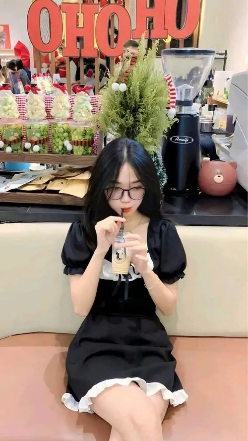 8 Cô nàng mặc váy đen ngồi trước nền bảng chữ sáng màu, tay giữ ly đồ uống nhỏ và cúi đầu nhẹ, kính mắt trong làm gương mặt thêm sáng, bức hình mang cảm giác xinh xắn, gọn ghẽ và rất hợp không khí vui tươi