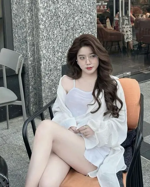 15 Hình cô gái ngồi trên ghế ngoài quán, váy trắng trễ vai làm nổi bật đôi chân và mái tóc xoăn bồng, bức này thiên về vẻ đẹp bí ẩn nhờ ánh nhìn mềm nhưng vẫn có thần thái và bố cục chừa khoảng trống đẹp