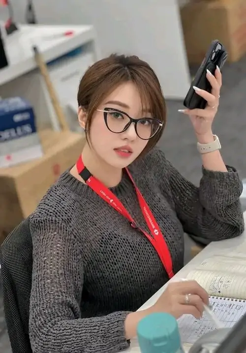 19 Hình girl tại bàn làm việc với kính gọng lớn, áo len ôm người và dây thẻ đỏ tạo chất công sở phá cách, khung hình trông sắc sảo hơn nhờ cánh tay nâng điện thoại và biểu cảm lạnh không dư thừa