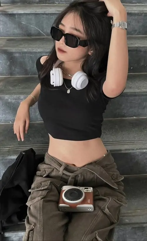 28 Cô gái ngồi trên bậc thang, đeo kính đen, mặc áo crop top cùng quần túi hộp, chiếc máy ảnh trước hông làm bố cục thêm nổi, tinh thần ảnh thời trang đường phố hiện rõ với dáng ngồi thoải mái và nắng gắt