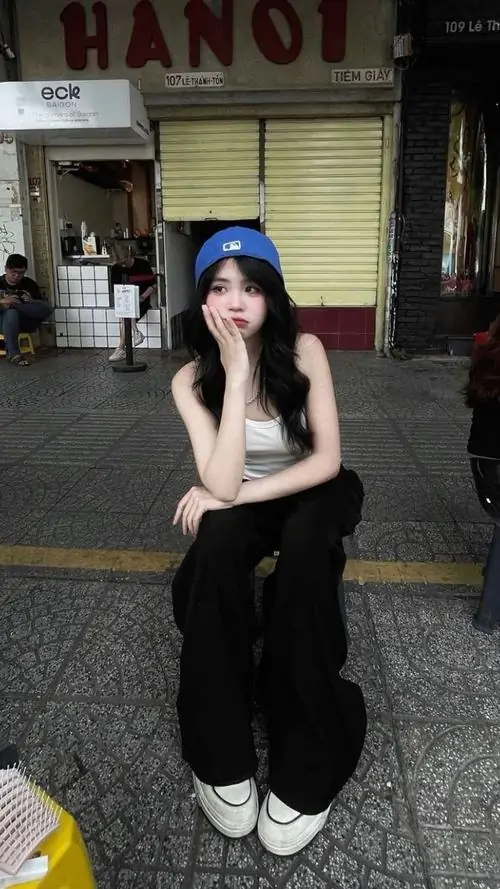 2 Cô nàng ngồi xổm ở lối đi vắng, đội mũ xanh và mặc đồ đen gọn gàng, pose chụm gối làm khung hình gợi phong cách cá tính mạnh, thêm chút khó gần từ cách nhìn thấp và nền phố cũ mờ phía sau