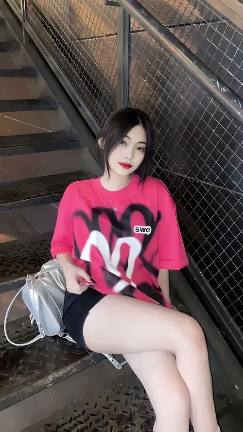 3 Hình gái tựa bên cầu thang sắt, áo hồng nổi bật trên nền tối, một tay giữ túi và chân xếp chéo nhẹ, tổng thể lên chất riêng nhờ quần short đen cùng cảm giác lạnh lùng từ góc chụp dọc khá gắt