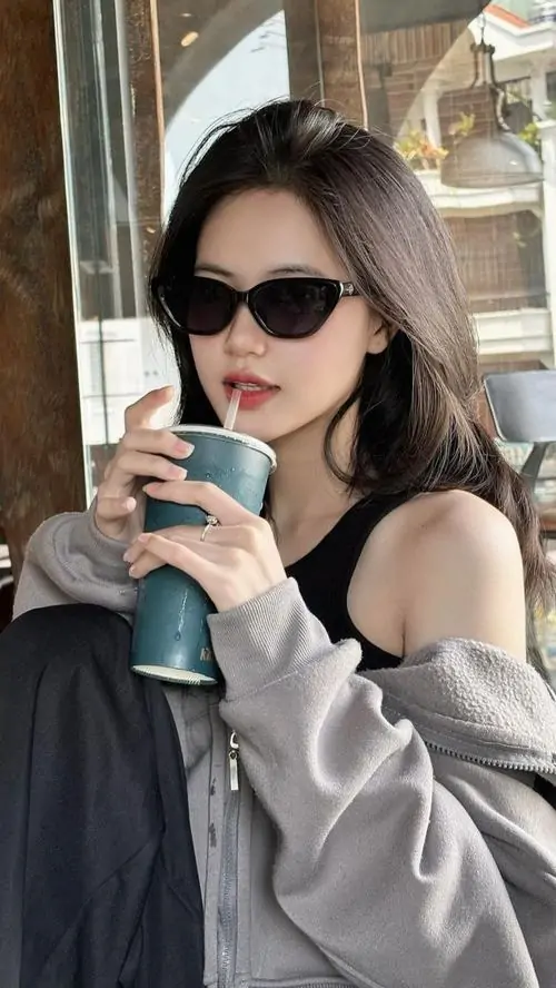 6 Cô gái đeo kính râm, ngồi trong quán kính sáng, tay cầm ly nước và khoác áo trễ vai màu xám, khung hình mang hơi hướng ảnh girl sang chảnh với outfit gọn, sắc sảo và ánh sáng dịu làm da mặt nổi bật