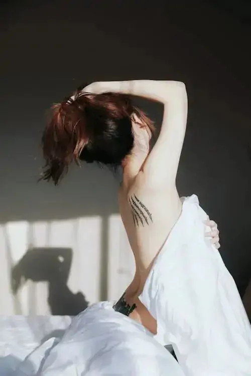 33 Dáng người xoay trong căn phòng sáng, váy trắng trượt khỏi vai để lộ phần lưng và cổ, nhiếp ảnh hình thể chọn bóng đổ trên tường như một lớp kể chuyện, khiến khung hình vừa mơ màng vừa có chiều sâu