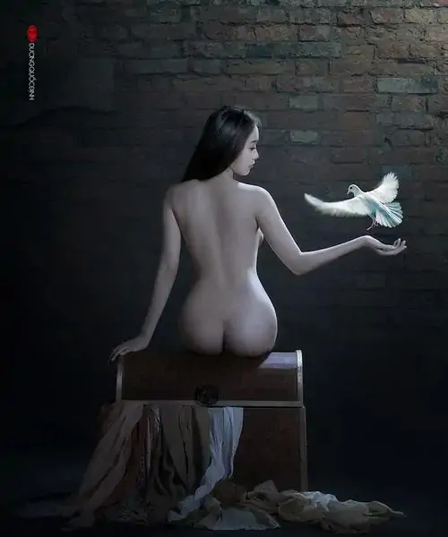 35 Người mẫu quay lưng ngồi trên bục gỗ trước tường gạch xám, một tay nâng nhành hoa trắng trong ánh đèn hẹp, bức ảnh nuy nghệ thuật làm rõ đường cong nghệ thuật và cảm giác cô tịch rất điện ảnh