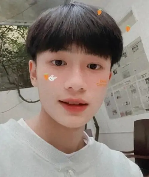 26 Nam sinh 2k12 áo đỏ đứng trong góc tối với tóc dựng nhẹ và khuôn mặt thanh, ảnh không quá cầu kỳ nhưng vẫn nổi bật nhờ nét non trẻ cùng sắc đỏ bắt mắt, nhìn vào thấy rõ chất Gen Alpha hồn nhiên