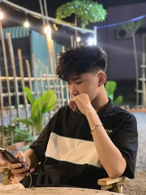 40 Nam sinh áo trắng cùng cà vạt ngồi cạnh chậu cây, cúi mặt vào lòng bàn tay như đang giấu tâm trạng, phần tóc buông che thêm mắt nên tổng thể vừa đẹp vừa trầm, rất hợp mood ảnh chân dung tối màu