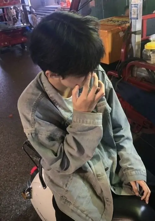 24 Một chàng trai mặc áo khoác xanh xám ngồi bên máy trò chơi, dùng tay và ống tay che gần hết miệng khi quay mặt nghiêng xuống, màu ảnh mờ lạnh khiến tổng thể dễ thương mà vẫn giữ vẻ xa cách