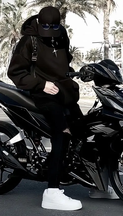 2 Chàng nam sinh đồ đen ngồi trên xe máy, trùm mũ kín và kéo khẩu trang cao che mặt, thân người co gọn giữa nền đường sáng nhạt, tạo cảm giác hấp dẫn, cá tính và rất hợp phong cách đường phố về đêm