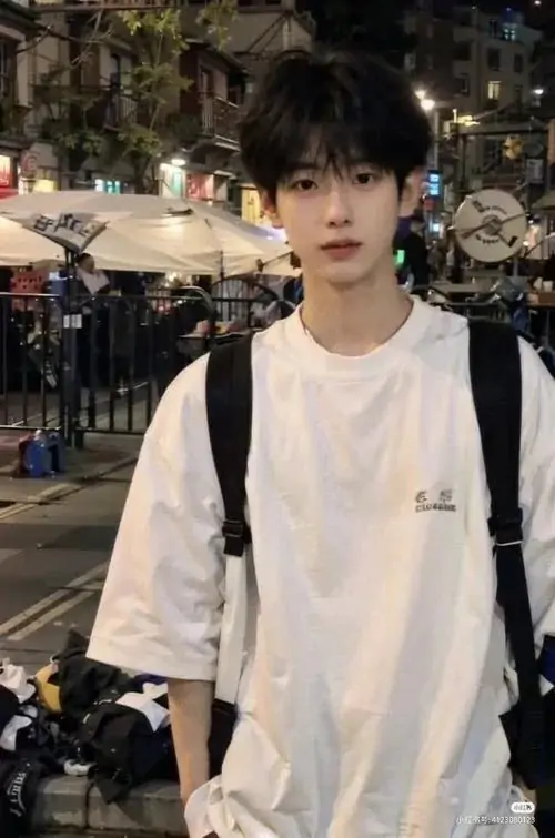 26 Chàng trai mặc áo sáng đứng trong khu phố đông người với dây đeo vai đen tạo nhịp tương phản rõ ràng giữa nền xô bồ và chủ thể tĩnh bức ảnh hợp kiểu street style nam nhờ dáng thẳng và nét mặt chắc