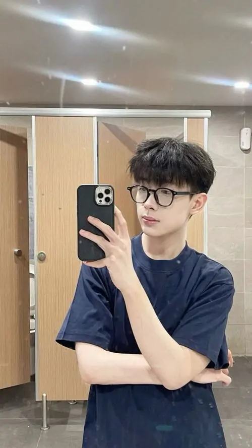 16 Hình soái ca đeo kính chụp gương trong phòng vệ sinh, chàng trai mặc áo tối giản và cầm điện thoại che nửa mặt, tư thế đứng thẳng giúp tỷ lệ cân đối, bầu không khí trẻ trung và có gu, hợp kiểu ảnh trai đẹp đeo kính đời thường