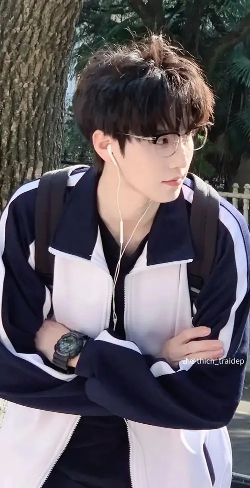 1 Ảnh trai đẹp đeo kính với áo khoác thể thao tông đen trắng, chàng trai khoanh tay đứng ngoài trời, nhìn nghiêng đầy điềm tĩnh, gọng kính mảnh làm khuôn mặt sáng hơn, tổng thể gợi cảm giác bí ẩn mà vẫn gần gũi trong khung hình dọc