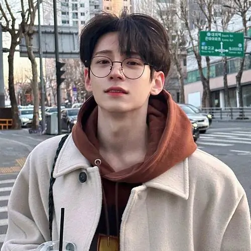 18 Ảnh trai đẹp đeo kính giữa phố mùa lạnh, chàng trai mặc hoodie nâu và khoác áo sáng màu, đứng thả lỏng trước hàng cây trơ lá, màu ảnh trung tính làm khung hình rất điện ảnh, gợi phong cách lịch lãm pha chút trầm lắng