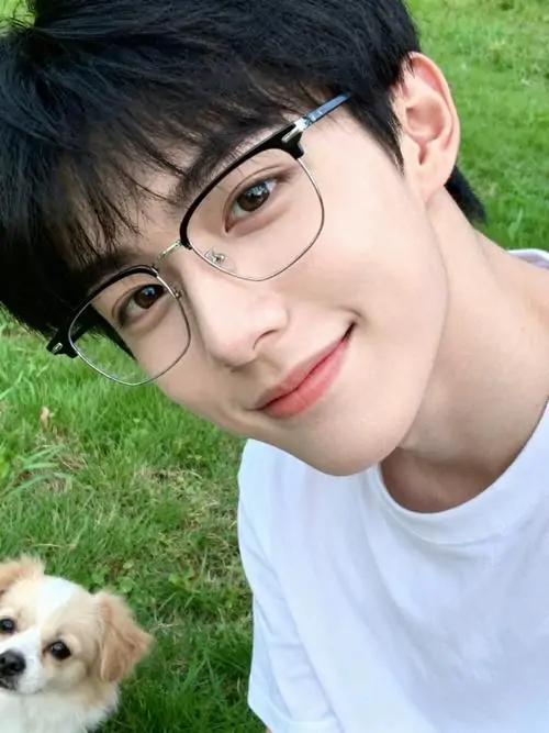 23 Ảnh chàng trai đeo kính ngoài cỏ xanh với áo trắng đơn giản, chàng trai nghiêng đầu cười nhẹ khi chú chó ló vào góc khung hình, ánh sáng tự nhiên làm da trông mềm, bức ảnh đem lại cảm giác trong trẻo và gần gũi