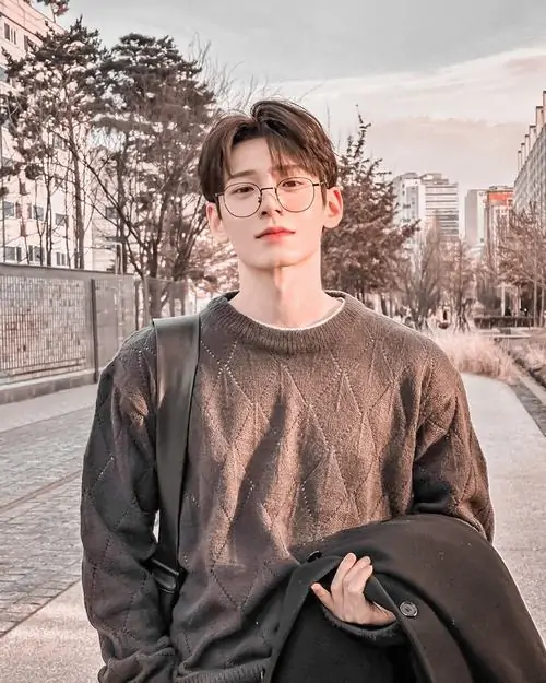 2 Chàng trai đứng giữa con đường vắng, mặc áo len nâu và cầm hờ áo khoác, mái tóc bồng nhẹ ôm trán, kiểu ảnh nam đẹp đeo kính mang vẻ thư sinh, ánh sáng lạnh ngoài trời giúp tổng thể thêm rõ nét và dễ mến