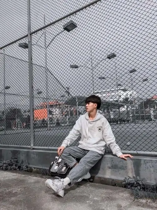 1 Ảnh trai đẹp k8 ngồi tựa bên lưới sân bóng, áo hoodie sáng màu và quần xám tạo cảm giác trẻ trung, dáng thả lỏng giữa khung nền gần như đơn sắc làm bật khí chất học đường và nét ngại ngùng rất vừa mắt