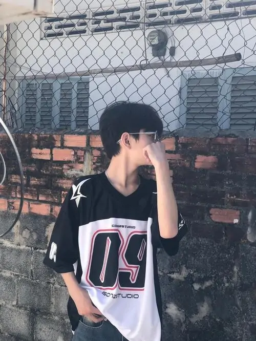 10 Chàng trai 2k8 tựa bên tường gạch, mặc áo thể thao rộng có số lớn phía trước và đưa tay chạm mặt, tổng thể đậm phong cách boy phố, nhìn qua đã thấy cá tính mạnh nhưng vẫn giữ được nét sáng sủa