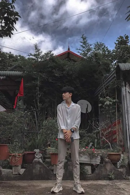 12 Bạn nam sinh 2008 đứng giữa khu vườn có chùa và mây xám phía xa, áo sáng cùng quần tối tạo độ cân bằng dễ nhìn, bức ảnh mang cảm giác thanh lịch, hợp nhóm hot boy học đường theo hướng trầm ổn