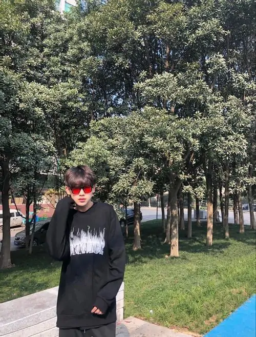 2 Cậu bạn 2k8 đứng trên bãi cỏ cạnh hàng cây, mặc áo tối màu và đeo kính đỏ tạo điểm nhấn lạ mắt, khung hình ngoài trời gợi vẻ năng động, hợp tinh thần đời thường của ảnh nam sinh điển trai đang được yêu thích