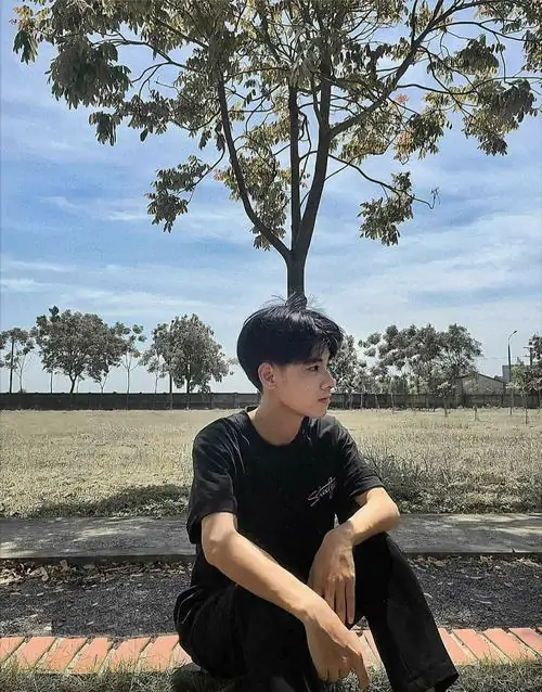 4 Bạn nam sinh ngồi dưới tán cây giữa nền trời xanh và bãi đất trống, áo đen làm tổng thể trầm hơn, góc chụp xa giữ cảm giác bí ẩn, rất hợp kiểu ảnh trai đẹp góc nghiêng mang chất thanh xuân yên yên